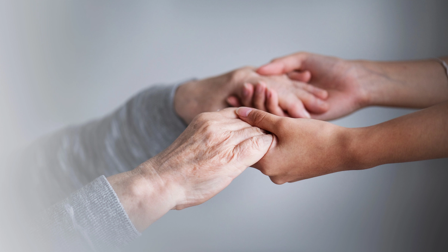 Custodia24 – 24-Stunden-Betreuungskräfte – Die Hände einer älteren Person werden sanft von den Händen einer jüngeren Person vor grauem Hintergrund gehalten